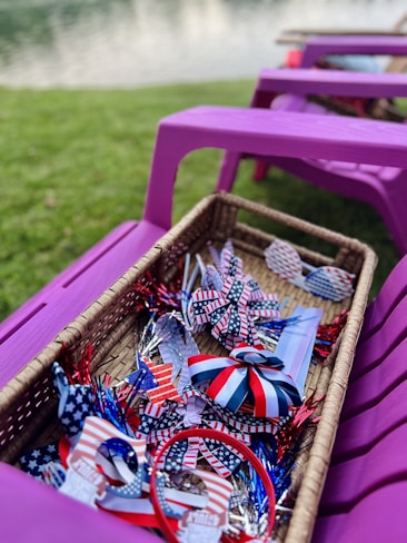 A wicker basket filled with patriotic-themed items, including ribbons and decorations in red, white, and blue with star patterns. The basket is placed on a purple outdoor chair with grass and a body of water in the background.
