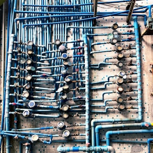 An intricate network of blue pipes and valves is mounted against a tiled wall. Numerous pressure gauges are attached at intervals, providing a detailed and complex arrangement. Each pipe is carefully connected, and there are various fittings and connections visible.