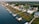 A coastal residential area features houses lined up along the water with private docks extending into the waterway. The homes are surrounded by lush greenery, and a road runs parallel to the shoreline.