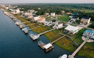 A coastal residential area features houses lined up along the water with private docks extending into the waterway. The homes are surrounded by lush greenery, and a road runs parallel to the shoreline.