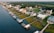 A coastal residential area features houses lined up along the water with private docks extending into the waterway. The homes are surrounded by lush greenery, and a road runs parallel to the shoreline.