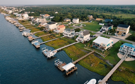 A coastal residential area features houses lined up along the water with private docks extending into the waterway. The homes are surrounded by lush greenery, and a road runs parallel to the shoreline.