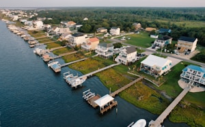 A coastal residential area features houses lined up along the water with private docks extending into the waterway. The homes are surrounded by lush greenery, and a road runs parallel to the shoreline.