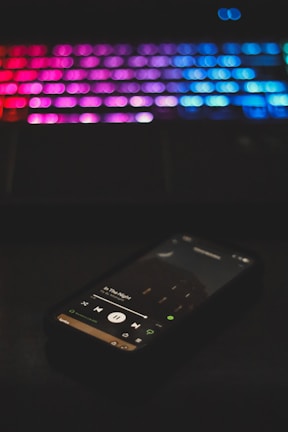 A smartphone displaying a music app playing a song titled 'In The Night' is in the foreground. The background features a blurred keyboard with colorful LED backlighting, showcasing shades of pink, purple, and blue.
