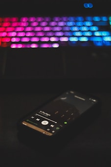 A smartphone displaying a music app playing a song titled 'In The Night' is in the foreground. The background features a blurred keyboard with colorful LED backlighting, showcasing shades of pink, purple, and blue.