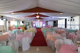 An elegantly decorated event hall with neatly arranged round tables covered in pastel-colored tablecloths and white chairs. A red carpet leads down the center aisle. The space is well-lit with chandeliers and spotlights, and floral centerpieces adorn each table.
