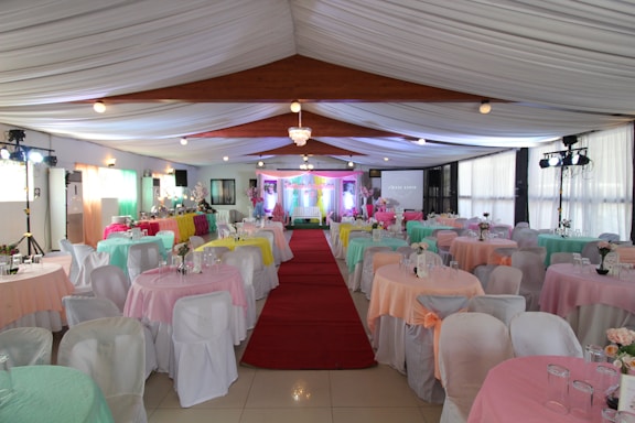 An elegantly decorated event hall with neatly arranged round tables covered in pastel-colored tablecloths and white chairs. A red carpet leads down the center aisle. The space is well-lit with chandeliers and spotlights, and floral centerpieces adorn each table.