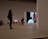 A modern gallery space featuring various art pieces on display. A large video screen shows a close-up image of an older man with a worn expression, wearing a hat and a blue shirt. Two people are in the gallery, one person standing and looking at their phone while another is walking with hands in pockets. Photographs are hung on the wall in the background, depicting outdoor scenes.