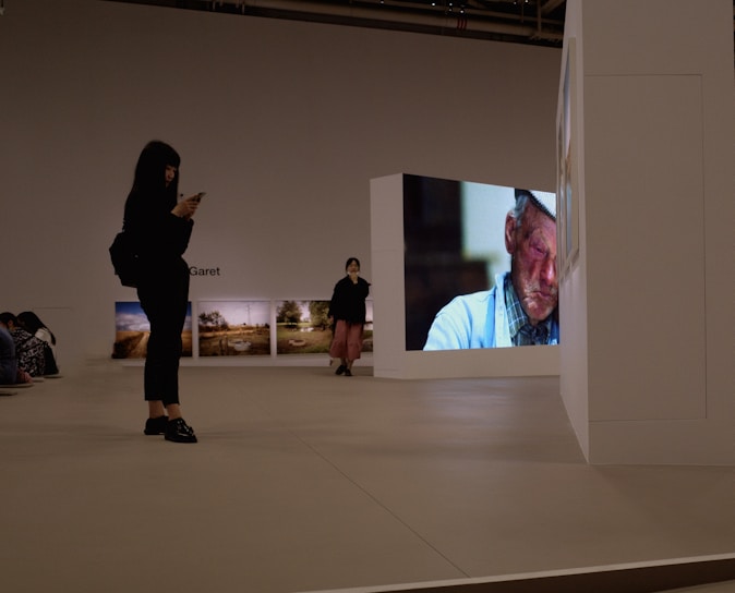 A modern gallery space featuring various art pieces on display. A large video screen shows a close-up image of an older man with a worn expression, wearing a hat and a blue shirt. Two people are in the gallery, one person standing and looking at their phone while another is walking with hands in pockets. Photographs are hung on the wall in the background, depicting outdoor scenes.