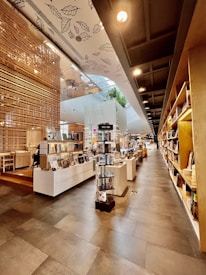 A spacious and modern bookstore with a well-organized layout. There are shelves filled with books along the right side of the image, and display tables in the center featuring various items such as books and magazines. The space has a high ceiling with decorative leaf designs, and there is a wooden partition on the left side of the image. Natural light filters through large windows and a skylight, enhancing the open and airy atmosphere. There are plants visible in the background, adding a touch of greenery to the space.