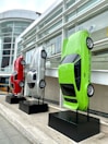Three cars are mounted vertically on black stands outside a modern building with large glass windows. The vehicles are displayed with their front ends facing upward. They are red, silver, and neon green in color and are positioned side by side on a stone platform.