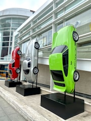 Three cars are mounted vertically on black stands outside a modern building with large glass windows. The vehicles are displayed with their front ends facing upward. They are red, silver, and neon green in color and are positioned side by side on a stone platform.