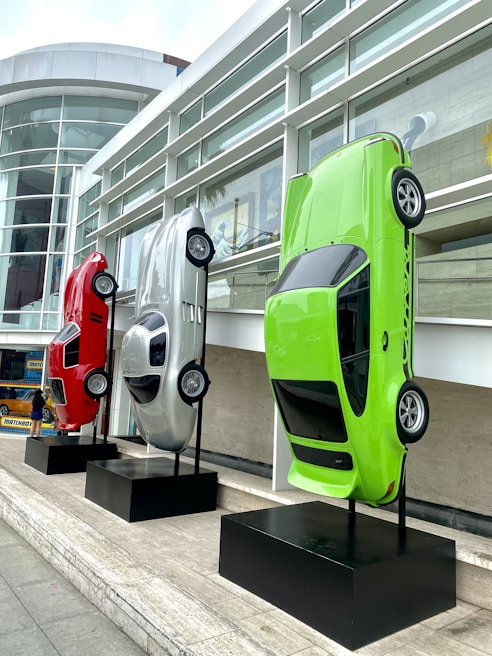Three cars are mounted vertically on black stands outside a modern building with large glass windows. The vehicles are displayed with their front ends facing upward. They are red, silver, and neon green in color and are positioned side by side on a stone platform.