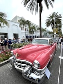 A classic pink Cadillac with chrome detailing is prominently displayed on a street lined with palm trees and a tall, white building in the background. Several people are gathered around, some observing the car and others strolling along. The setting appears to be a car show or exhibition, with a sign in front of the vehicle indicating details about the Cadillac.