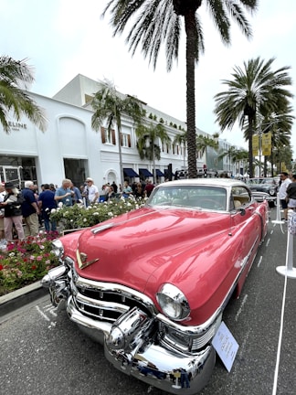 A classic pink Cadillac with chrome detailing is prominently displayed on a street lined with palm trees and a tall, white building in the background. Several people are gathered around, some observing the car and others strolling along. The setting appears to be a car show or exhibition, with a sign in front of the vehicle indicating details about the Cadillac.
