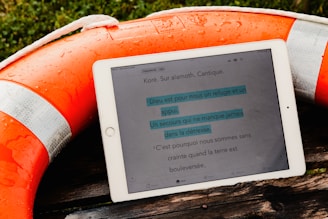 A tablet displaying text in French is resting on a wooden surface next to an orange life buoy with silver reflective bands. The life buoy is wet, indicating recent exposure to water. The text on the tablet seems to be from a religious scripture or song, possibly a Psalm. The scene is set outdoors with some greenery in the background.