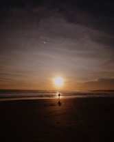 A silhouette of a person running along a beach, with a vast expanse of sand and the ocean reflecting the glowing light of a setting sun. The sky is painted with soft clouds, adding to the serene atmosphere of the scene.