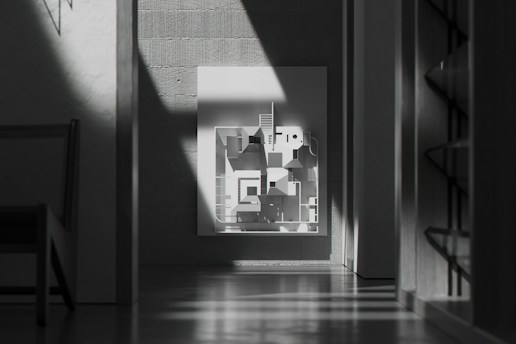A modern, minimalist interior features a monochrome geometric artwork on a wall. Sunlight casts dramatic shadows across the scene, highlighting the textures of the concrete walls. There is a chair partially visible on the left and shelving units on the right.