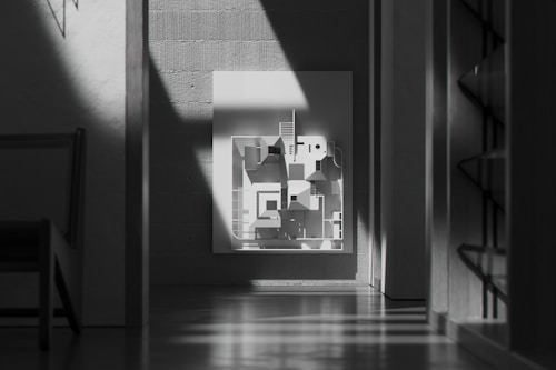 A modern, minimalist interior features a monochrome geometric artwork on a wall. Sunlight casts dramatic shadows across the scene, highlighting the textures of the concrete walls. There is a chair partially visible on the left and shelving units on the right.