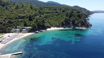 A picturesque coastal scene featuring a sandy beach lined with sun loungers and umbrellas. The beach is surrounded by lush, green hills and clear turquoise water. The landscape includes a mix of dense forest and rocky cliffs, with several small buildings nestled among the trees near the shore.