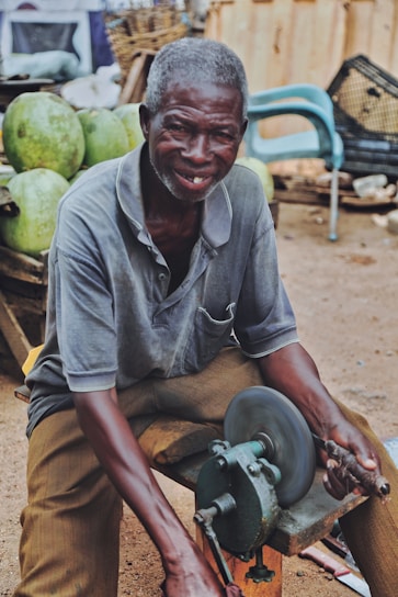 An elderly man with gray hair engages in a task using a manual grinding machine. He is smiling and appears to be enjoying his work. Behind him, there are several large green watermelons stacked, and various items are scattered in the background, including a blue plastic chair and wicker baskets.