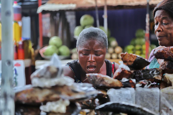 Two women are seen in a market setting, appearing to be observing or negotiating over various items laid out on a table in front of them. The background includes blurred stacks of fruits or vegetables, and colorful bottles on a shelf.