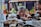 Two women are seen in a market setting, appearing to be observing or negotiating over various items laid out on a table in front of them. The background includes blurred stacks of fruits or vegetables, and colorful bottles on a shelf.