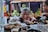 Two women are seen in a market setting, appearing to be observing or negotiating over various items laid out on a table in front of them. The background includes blurred stacks of fruits or vegetables, and colorful bottles on a shelf.