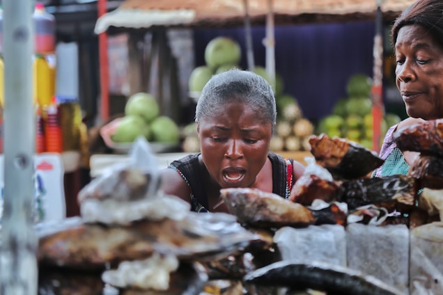 Two women are seen in a market setting, appearing to be observing or negotiating over various items laid out on a table in front of them. The background includes blurred stacks of fruits or vegetables, and colorful bottles on a shelf.