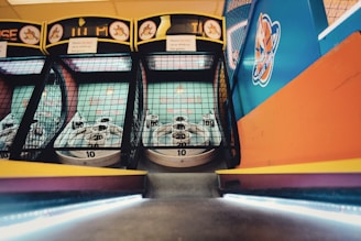 A row of skee-ball machines is positioned in an arcade setting. The skee-ball lanes are enclosed by mesh netting, and each lane has targets with point values labeled inside circular holes. The colorful design includes a mix of orange, blue, and yellow accents, and arcade game graphics are visible.