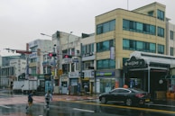 A street scene in an urban area with multiple small businesses featuring Korean signage. The buildings are several stories high with blue-tinted windows and visible signs for various shops. The wet pavement indicates recent rain, and a few pedestrians are crossing the street. A black sedan with a taxi sign on top is parked near the sidewalk.