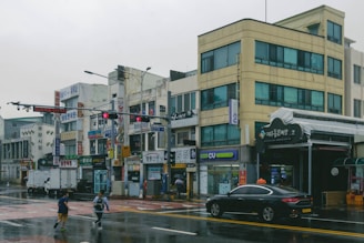 A street scene in an urban area with multiple small businesses featuring Korean signage. The buildings are several stories high with blue-tinted windows and visible signs for various shops. The wet pavement indicates recent rain, and a few pedestrians are crossing the street. A black sedan with a taxi sign on top is parked near the sidewalk.