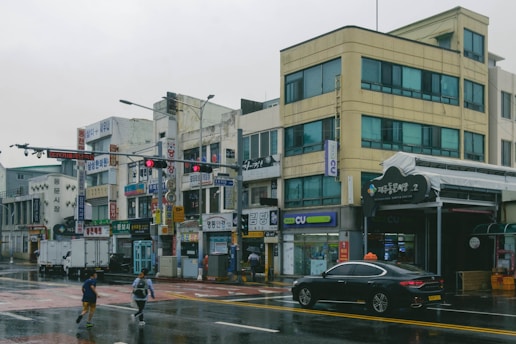 A street scene in an urban area with multiple small businesses featuring Korean signage. The buildings are several stories high with blue-tinted windows and visible signs for various shops. The wet pavement indicates recent rain, and a few pedestrians are crossing the street. A black sedan with a taxi sign on top is parked near the sidewalk.