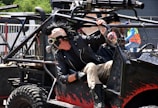 A person wearing a punk-inspired outfit with a spiked leather jacket and a mask featuring goggles is seated in a heavily customized vehicle. The vehicle is adorned with skull decorations and equipped with rugged tires. The scene suggests a post-apocalyptic or Mad Max-like theme.