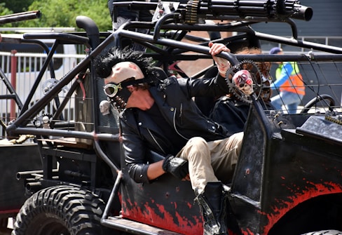 A person wearing a punk-inspired outfit with a spiked leather jacket and a mask featuring goggles is seated in a heavily customized vehicle. The vehicle is adorned with skull decorations and equipped with rugged tires. The scene suggests a post-apocalyptic or Mad Max-like theme.