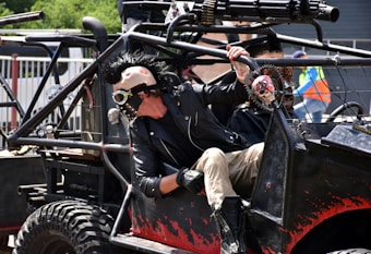 A person wearing a punk-inspired outfit with a spiked leather jacket and a mask featuring goggles is seated in a heavily customized vehicle. The vehicle is adorned with skull decorations and equipped with rugged tires. The scene suggests a post-apocalyptic or Mad Max-like theme.