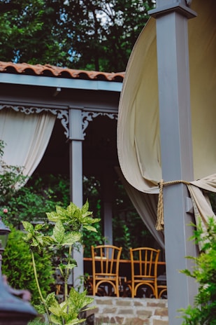 A serene outdoor setting with a wooden pergola adorned with decorative trim and cream-colored drapes. Beneath the pergola is a seating area with wooden chairs around a table. Lush greenery and variegated leaves beautify the foreground, while a garden background adds to the tranquil atmosphere.