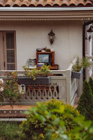 A cozy balcony setting featuring a small table with a classic television, various kitchen items, and plants. The balcony is adorned with ornate railings, and there is greenery in the foreground. A wall-mounted lamp and a traditional roof design add to the quaint charm.