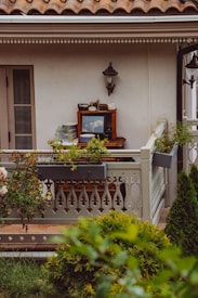 A cozy balcony setting featuring a small table with a classic television, various kitchen items, and plants. The balcony is adorned with ornate railings, and there is greenery in the foreground. A wall-mounted lamp and a traditional roof design add to the quaint charm.