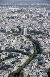 An aerial view of a densely packed urban area with a prominent historical monument at the center, surrounded by a network of streets and lush green trees. The architecture showcases a mix of classical and modern buildings with light-colored facades.