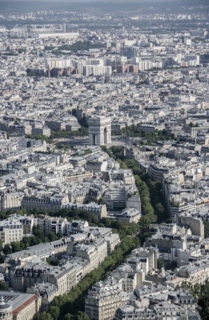 An aerial view of a densely packed urban area with a prominent historical monument at the center, surrounded by a network of streets and lush green trees. The architecture showcases a mix of classical and modern buildings with light-colored facades.