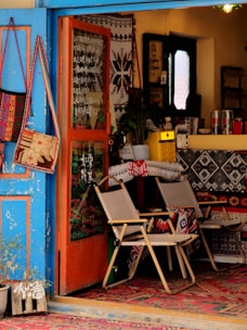 A cozy, eclectic shop interior featuring a mix of colorful, decorative textiles and furniture. The blue, open door invites you into a warm space filled with patterned rugs and chairs draped in textile designs. On the wall, bags hang as decorations alongside other woven art pieces. A counter at the back is partially visible, holding various items and a plant, contributing to a welcoming, artistic atmosphere.