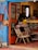 A cozy, eclectic shop interior featuring a mix of colorful, decorative textiles and furniture. The blue, open door invites you into a warm space filled with patterned rugs and chairs draped in textile designs. On the wall, bags hang as decorations alongside other woven art pieces. A counter at the back is partially visible, holding various items and a plant, contributing to a welcoming, artistic atmosphere.