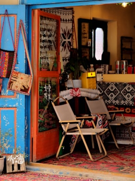 A cozy, eclectic shop interior featuring a mix of colorful, decorative textiles and furniture. The blue, open door invites you into a warm space filled with patterned rugs and chairs draped in textile designs. On the wall, bags hang as decorations alongside other woven art pieces. A counter at the back is partially visible, holding various items and a plant, contributing to a welcoming, artistic atmosphere.