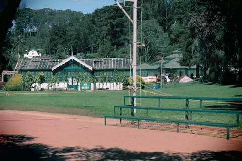A green building with a pitched roof surrounded by lush greenery and trees is prominently featured. The front of the building is adorned with plants and a 'No Entry' sign is visible. Open space with benches and a neatly maintained lawn stretches out in front of the building.