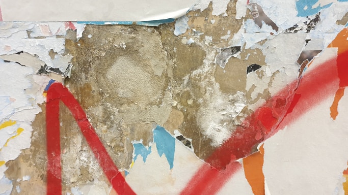 A textured surface with layers of peeling paint and paper, revealing a mix of beige and brown underneath. Broad red paint strokes create a dramatic effect against the worn backdrop. Light blue and orange fragments add contrast and visual interest.