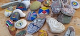 A collection of colorful, hand-painted stones arranged on a wooden surface. Each stone features unique designs, including abstract patterns, geometric shapes, a face, and nature-inspired motifs like trees and flowers. The stones vary in shape and size, showcasing an array of artistry and creativity in their decoration.