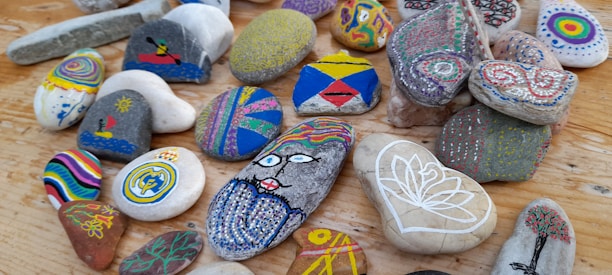 A collection of colorful, hand-painted stones arranged on a wooden surface. Each stone features unique designs, including abstract patterns, geometric shapes, a face, and nature-inspired motifs like trees and flowers. The stones vary in shape and size, showcasing an array of artistry and creativity in their decoration.