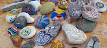 A collection of colorful, hand-painted stones arranged on a wooden surface. Each stone features unique designs, including abstract patterns, geometric shapes, a face, and nature-inspired motifs like trees and flowers. The stones vary in shape and size, showcasing an array of artistry and creativity in their decoration.