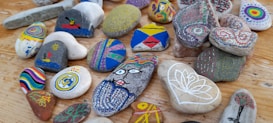 A collection of colorful, hand-painted stones arranged on a wooden surface. Each stone features unique designs, including abstract patterns, geometric shapes, a face, and nature-inspired motifs like trees and flowers. The stones vary in shape and size, showcasing an array of artistry and creativity in their decoration.